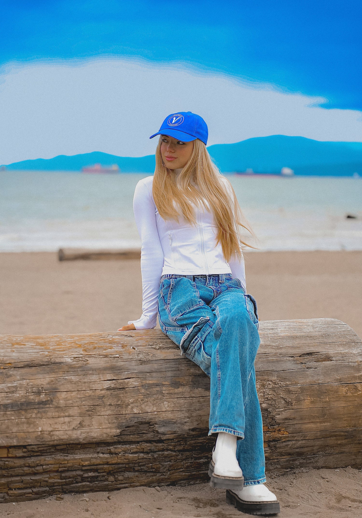 Female model sitting on a beach wearing the pacific and white Elation relaxed-fit hat by Vancouver Signature