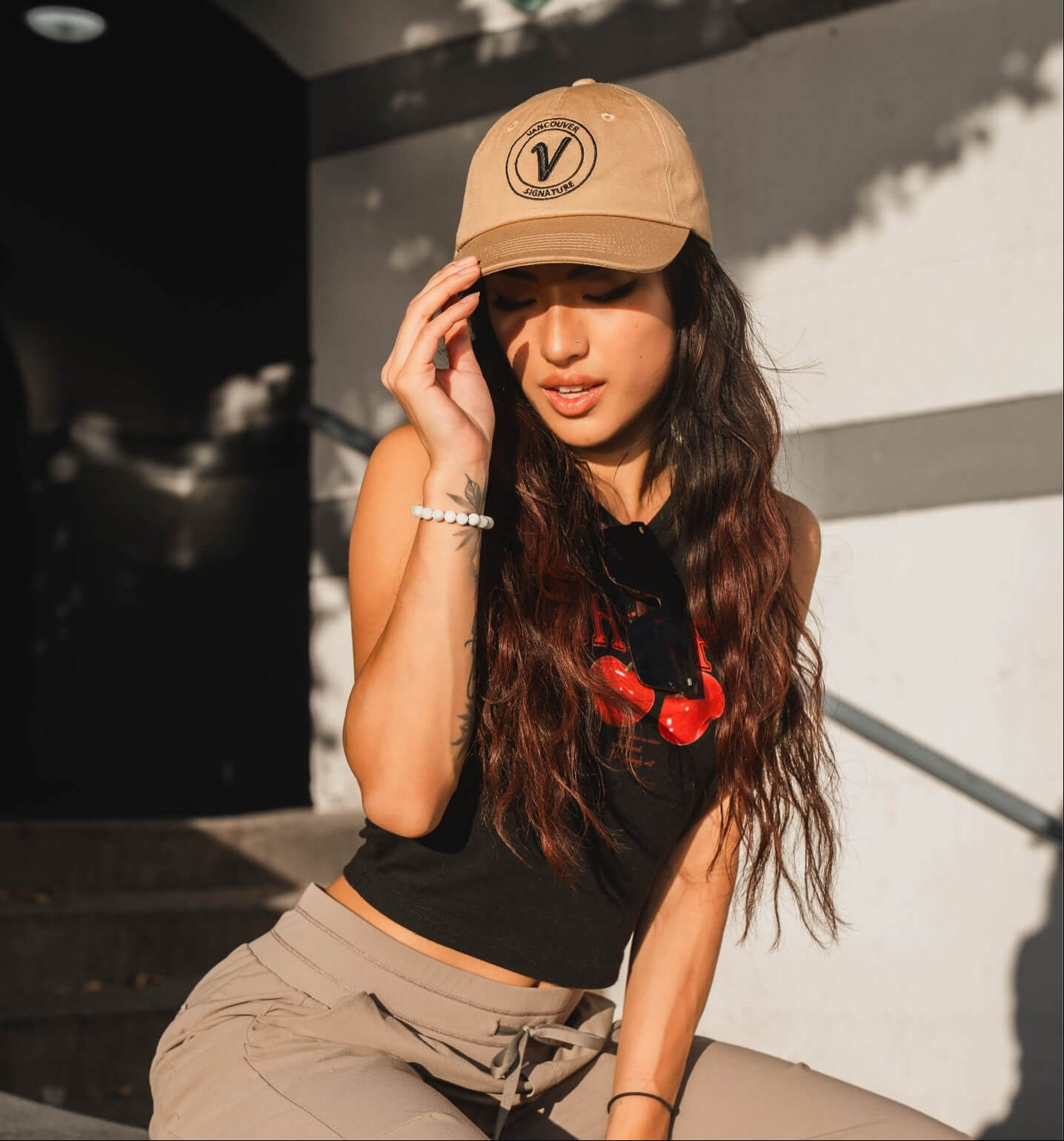 Female model wearing the sands and black Elation relaxed-fit hat by Vancouver Signature outdoors in a sunny setting.