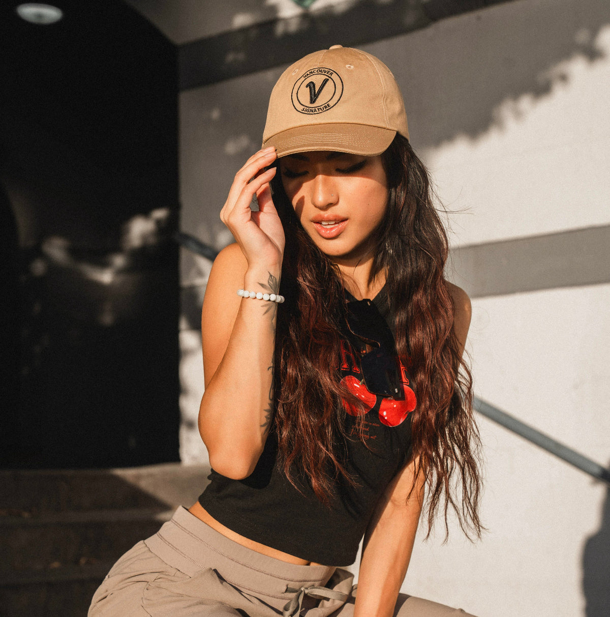 Female model wearing the sands and black Elation relaxed-fit hat by Vancouver Signature outdoors in a sunny setting.