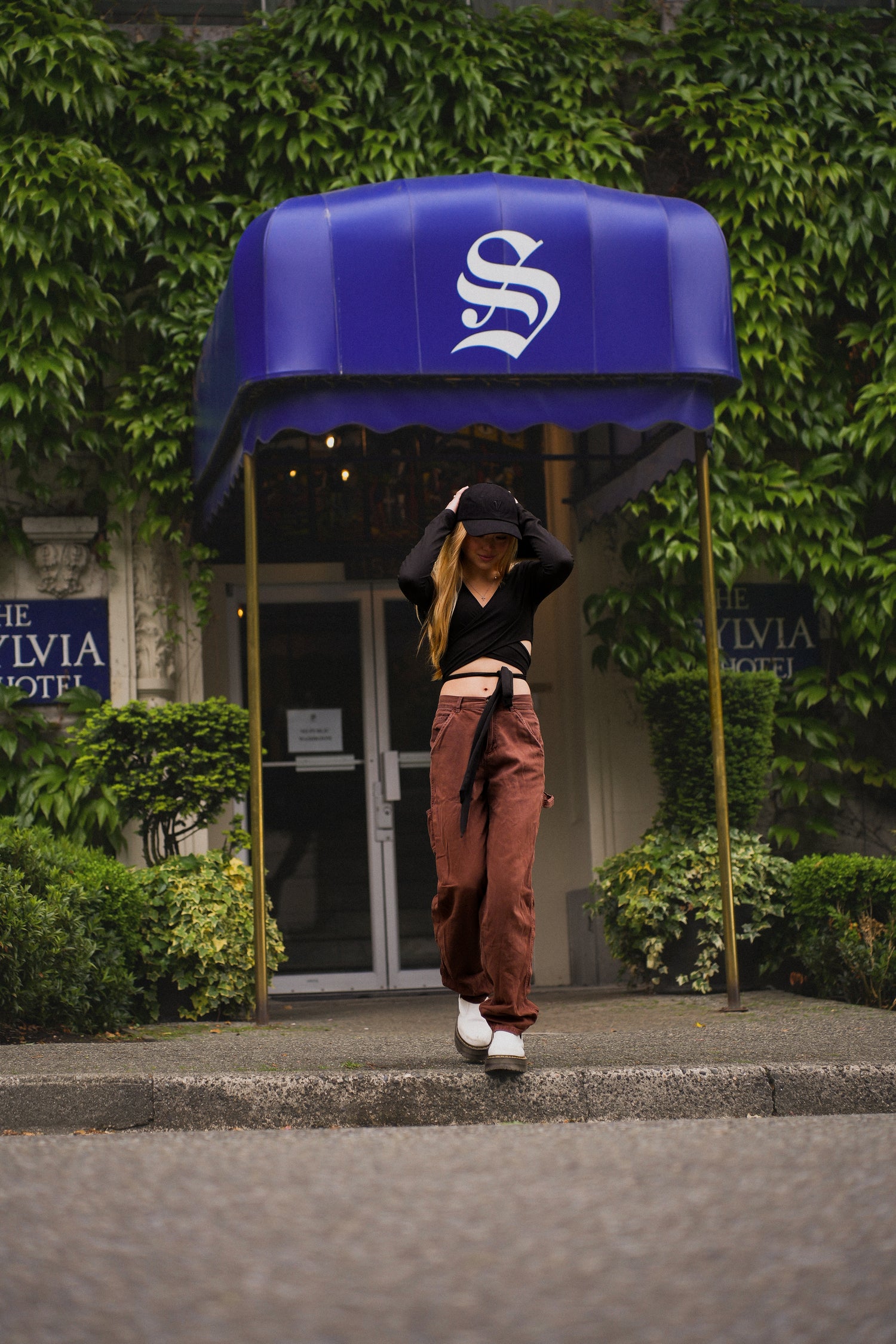 Female model crossing a street in front of a blue sign wearing the Elation Hat in black from Vancouver Signature