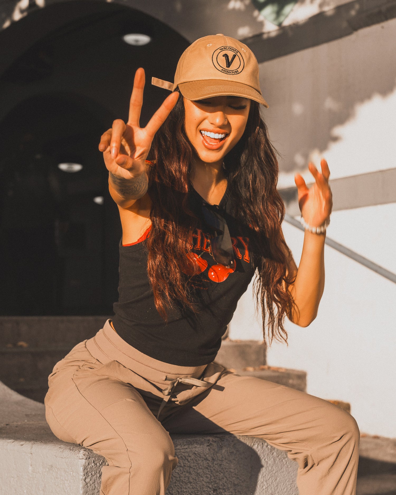 Female model sitting on a ledge showing the peace sign and wearing the Elation hat in sands and black by Vancouver Signature